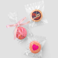 The Custom Single Macaron Bag by Jenna Rae Cakes includes three individually wrapped round macarons: one with a pink heart, one with pink icing and sprinkles, and one with an orange Jets logo on a blue background.