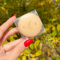 A hand with bright red nails holds a Custom Single Macaron Box by Jenna Rae Cakes in beige, with gold C & E lettering. The background features blurred yellow-green leaves.
