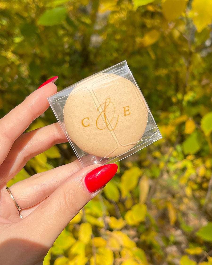 A hand with bright red nails holds a Custom Single Macaron Box by Jenna Rae Cakes in beige, with gold C & E lettering. The background features blurred yellow-green leaves.