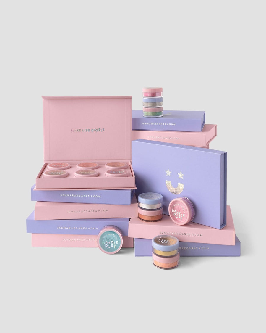 Pastel pink and lavender Dazzle Dust Boxes and jars by Jenna Rae Cakes, some open to reveal shimmer powders. The minimalist packaging features the phrase MAKE LIFE DAZZLE.