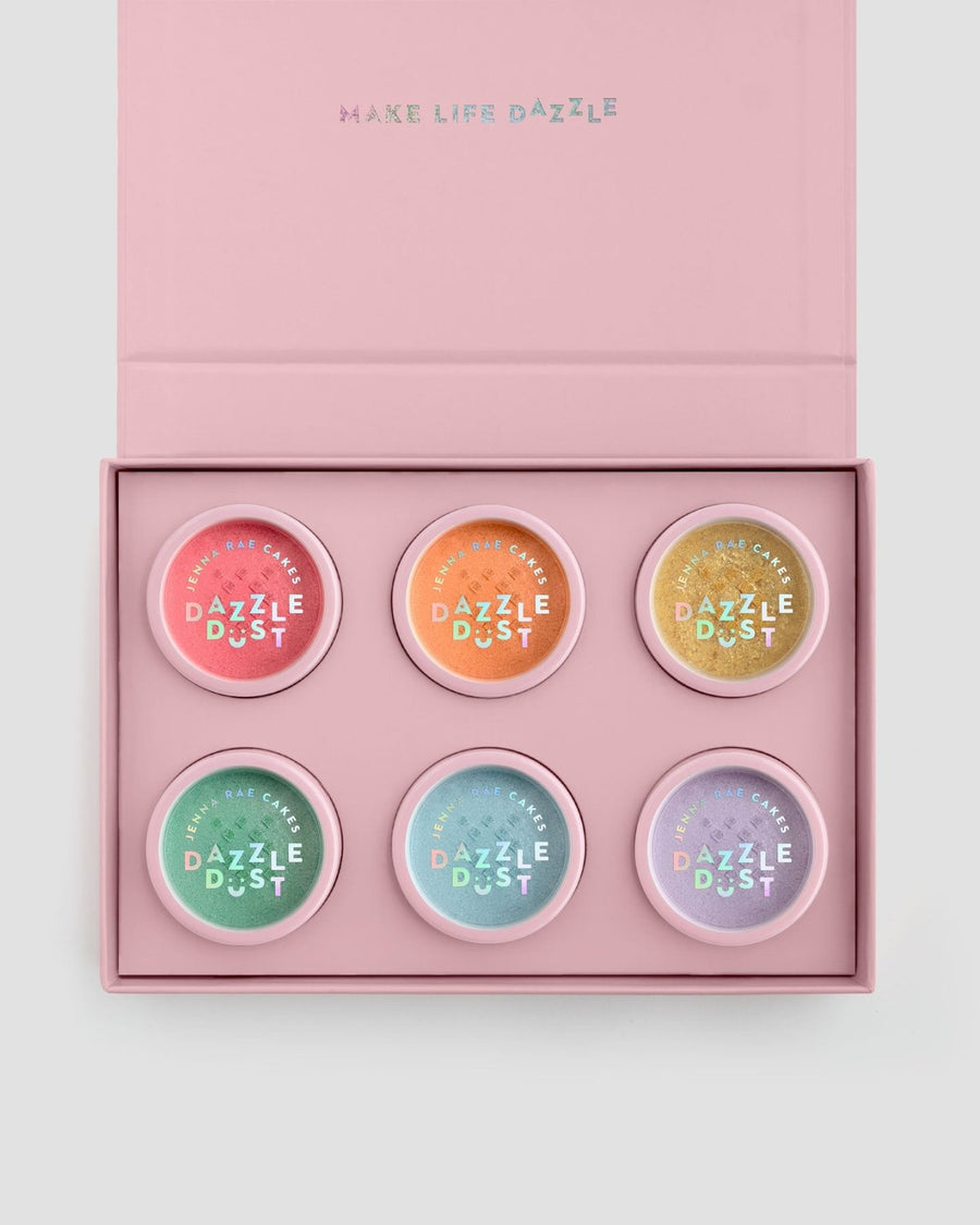The Dazzle Dust Box by Jenna Rae Cakes is a pink box with “MAKE LIFE DAZZLE” on the lid, featuring six pastel-colored circular tins labeled “DAZZLE DUST,” arranged in two neat rows inside.