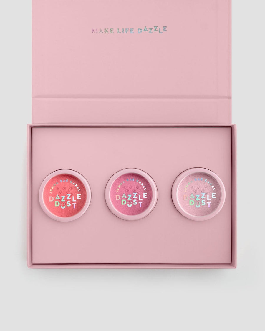 The Jenna Rae Cakes Dazzle Dust Box is an open pink box with “MAKE LIFE DAZZLE” inside the lid, holding three round “Dazzle Dust” makeup containers in varying pink shades.
