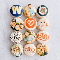 A set of twelve macarons arranged in a 3x4 grid, featuring custom-printed images and logos, is showcased as part of the Dessert Tables collection by Jenna Rae Cakes.