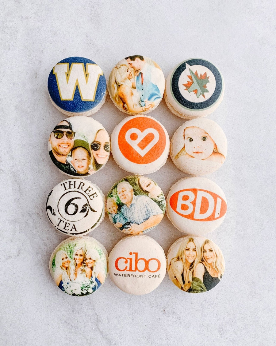 A set of twelve macarons arranged in a 3x4 grid, featuring custom-printed images and logos, is showcased as part of the Dessert Tables collection by Jenna Rae Cakes.