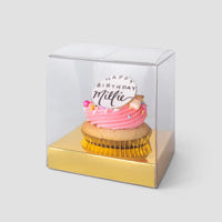A Jenna Rae Cakes Favours cupcake with pink frosting and a Happy Birthday Millie topper sits in a clear plastic box with a gold base against a light grey background.