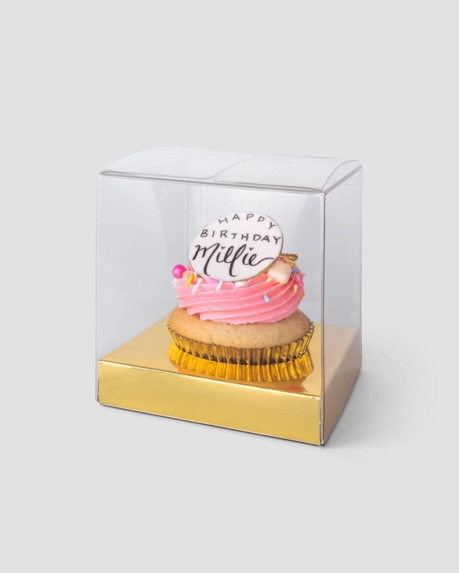 A Jenna Rae Cakes Favours cupcake with pink frosting and a Happy Birthday Millie topper sits in a clear plastic box with a gold base against a light grey background.