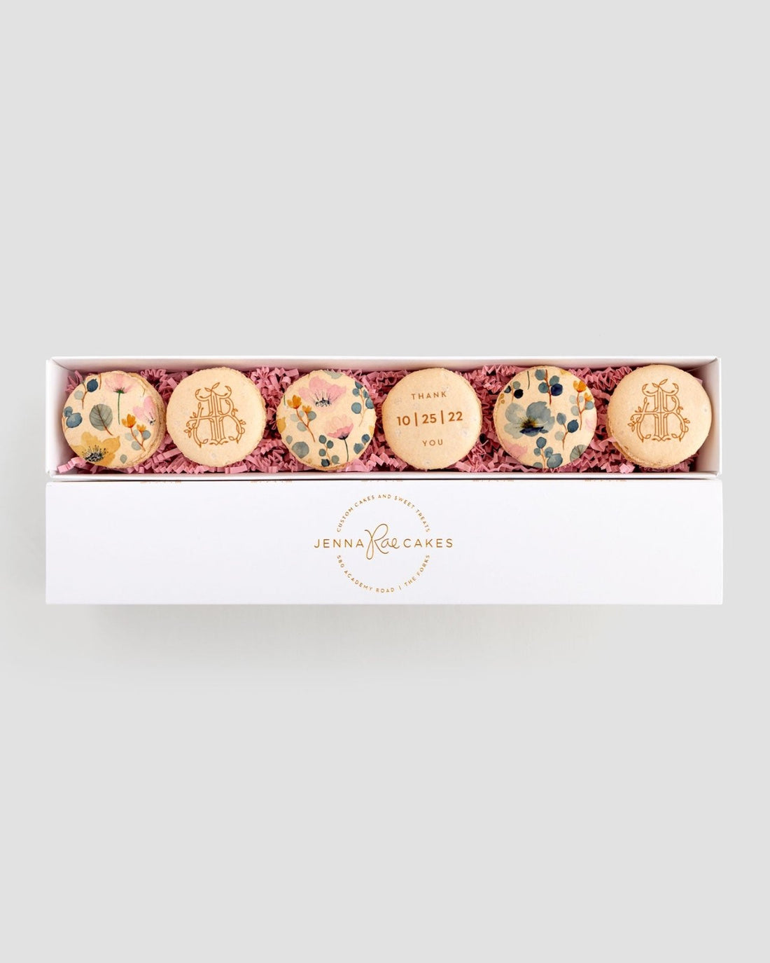 A white box labeled Jenna Rae Cakes contains six Favours macarons with floral designs and text, nestled in pink shredded paper. The center macaron features THANK YOU 10.15.22.