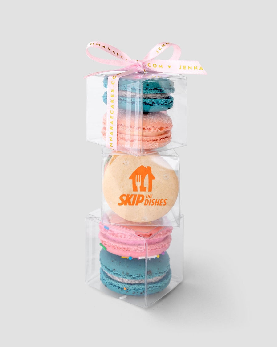 A stack of three clear Jenna Rae Cakes Favours boxes, each filled with colorful macarons and tied with a pink ribbon. The middle box features a macaron printed with the SkipTheDishes logo.