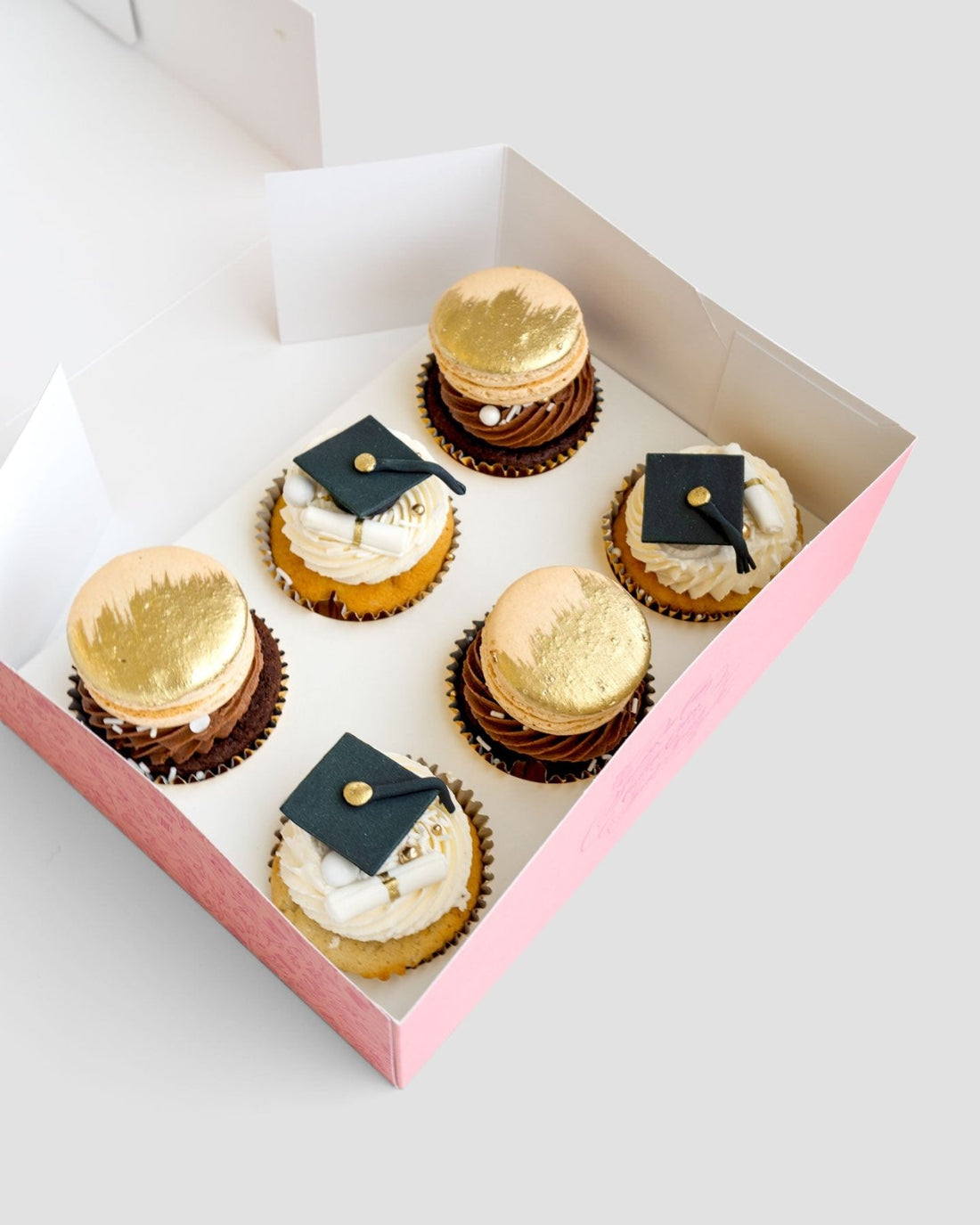 Grad Works Cupcakes by Jenna Rae Cakes: a pink box with six decorated cupcakes, three topped with gold macarons and three adorned with edible graduation caps and diplomas, arranged neatly on a light background.