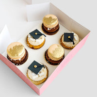 Grad Works Cupcakes by Jenna Rae Cakes: a pink box with six decorated cupcakes, three topped with gold macarons and three adorned with edible graduation caps and diplomas, arranged neatly on a light background.