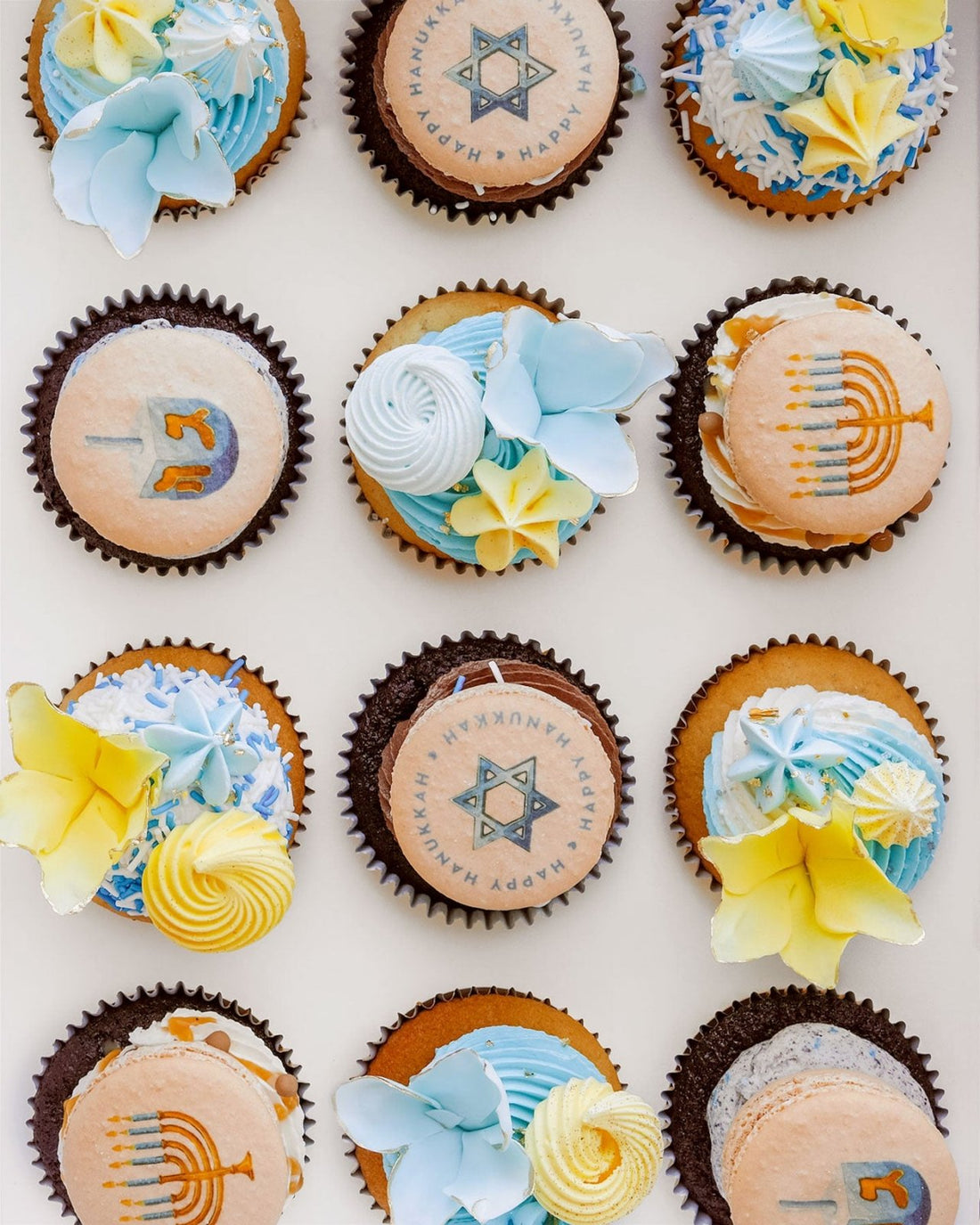 Hanukkah Works Cupcakes by Jenna Rae Cakes are decorated with blue, yellow, and white flowers, menorahs, dreidels, and Stars of David—perfectly arranged for a festive Hanukkah celebration.