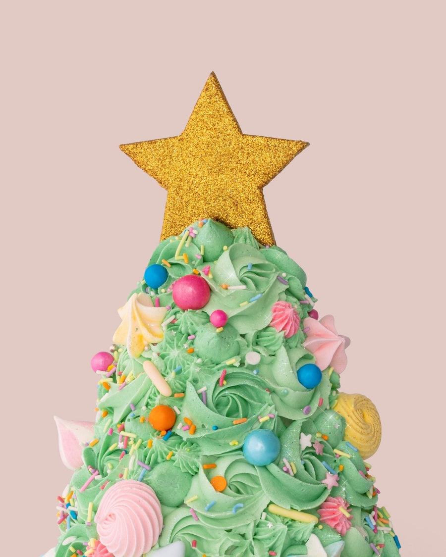 The Christmas Tree Cake by Jenna Rae Cakes features green frosting, colorful candy ornaments, and a glittery gold star on top, set against a plain light pink background.