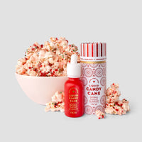 A bowl of popcorn with red and white sprinkles sits beside a small red bottle and a tall white tin labeled Liquid Candy Cane by Jenna Rae Cakes, all placed on a plain light gray background.