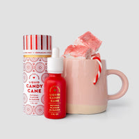 A pink mug with pink and white marshmallows and a small candy cane sits beside a Jenna Rae Cakes Liquid Candy Cane dropper bottle and matching cylindrical container.