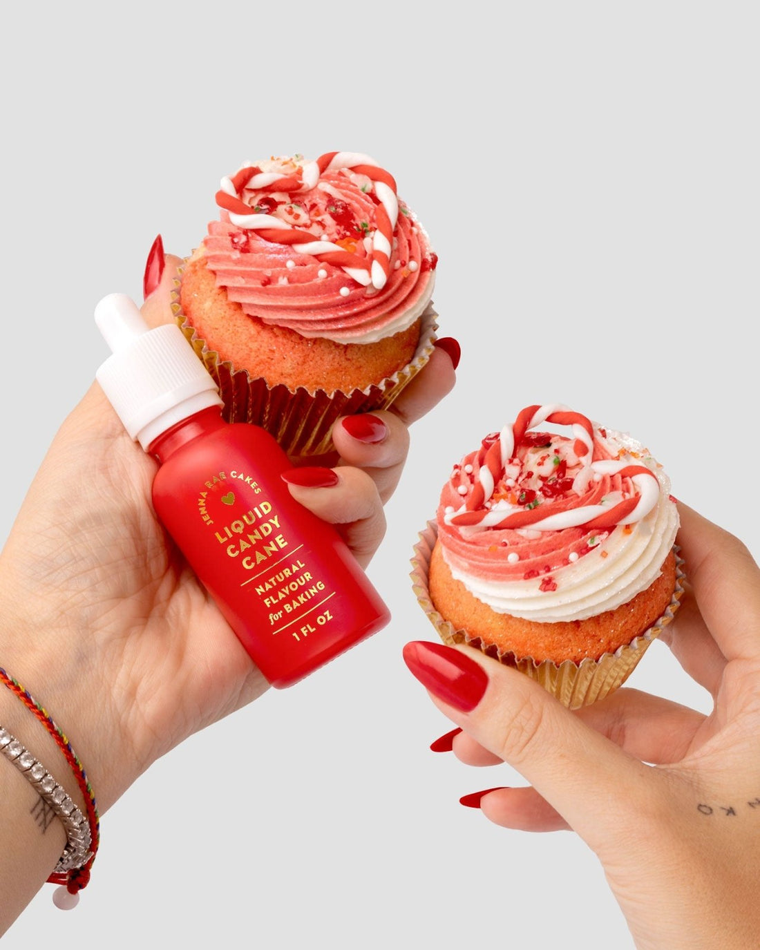 Two hands with red nails hold frosted red and white cupcakes; one hand also holds a Jenna Rae Cakes Liquid Candy Cane dropper bottle, showing it can be used for decorating or flavoring desserts.