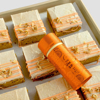 A baking sheet with frosted pumpkin spice dessert bars, topped with orange drizzle and chopped nuts, features a Jenna Rae Cakes Liquid Pumpkin Spice container resting diagonally on top of the bars.