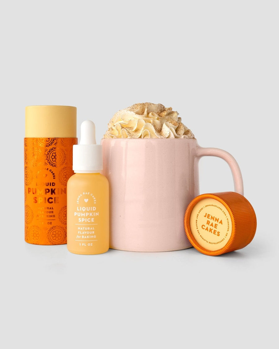 A pink mug with whipped cream sits beside orange and yellow containers and a Jenna Rae Cakes Liquid Pumpkin Spice dropper bottle, all set against a light gray background.