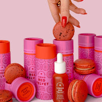A hand with red nails holds a red macaron above pink tubes of Jenna Rae Cakes Liquid Red Velvet and a small dropper bottle, surrounded by more red macarons and product containers.