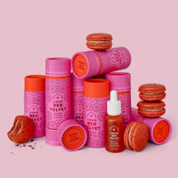 Jenna Rae Cakes Liquid Red Velvet tubes are stacked with whole and broken red velvet macarons and a dropper bottle, all on a pink background.