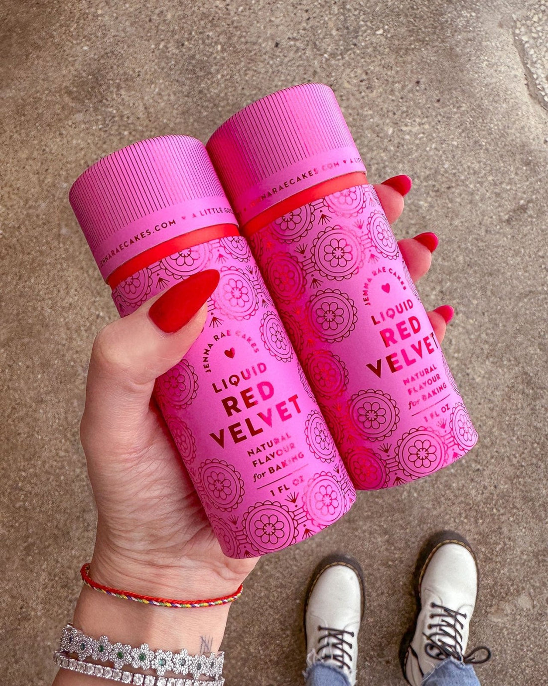 A hand with red-painted nails holds two pink Jenna Rae Cakes Liquid Red Velvet bottles against a brown textured background. White shoes and part of a bracelet are visible at the bottom of the image.