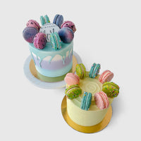 The Macaron Crown Cake by Jenna Rae Cakes features two cakes topped with colorful macarons, one with pastel blue and purple icing and white drip, the other with light yellow icing. Both sit on gold bases against a plain white background.