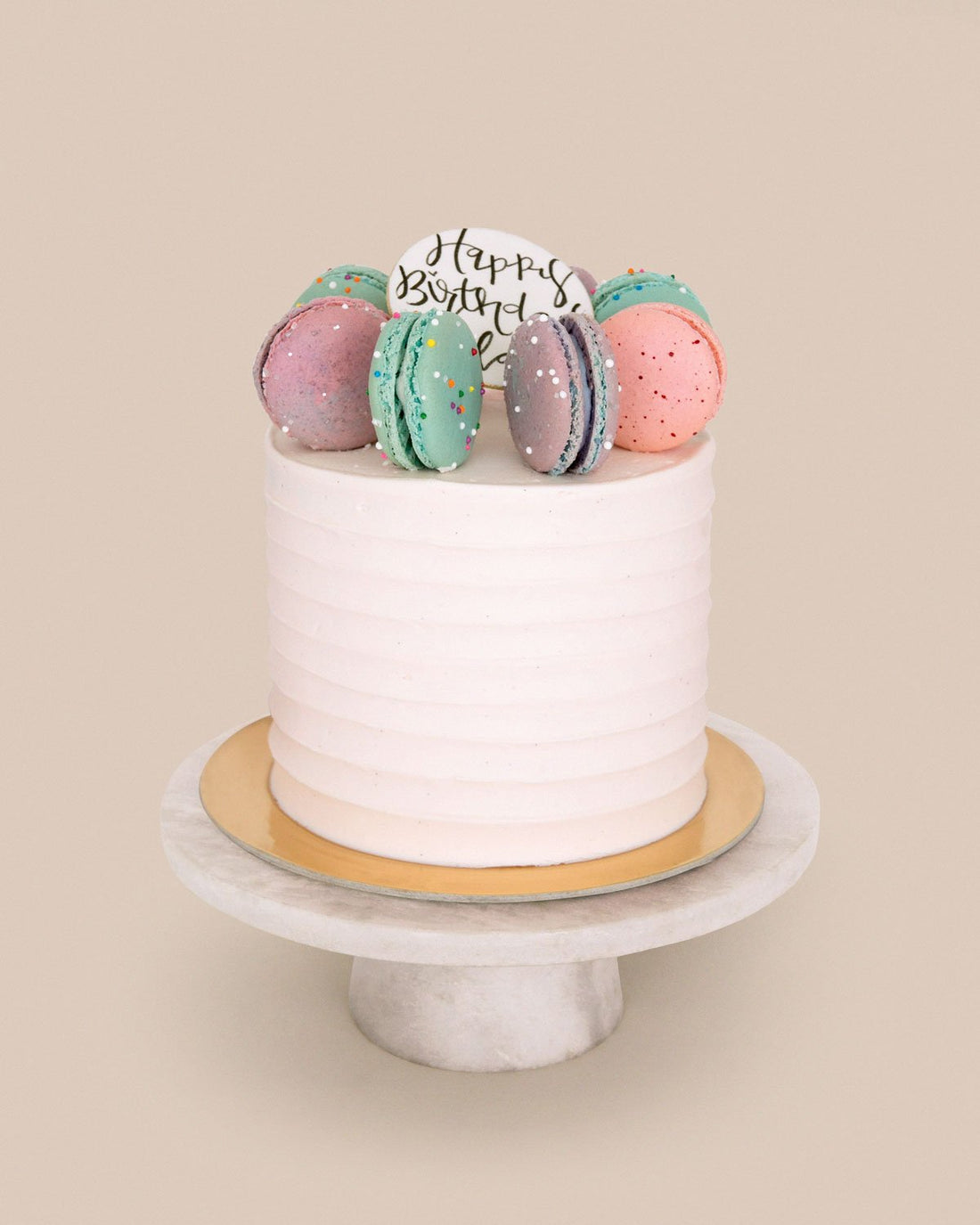 The Macaron Crown Cake by Jenna Rae Cakes features white frosting, colorful macarons, and a Happy Birthday sign, displayed on a round marble cake stand against a beige background.