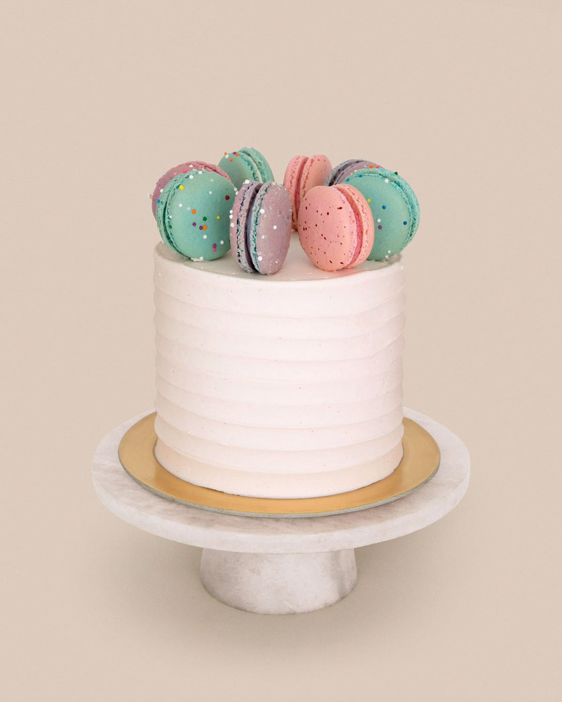 The Macaron Crown Cake by Jenna Rae Cakes features a white frosted finish topped with colorful macarons, displayed on a gold cake board and white marble stand, set against a plain beige background.
