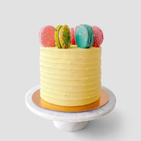 The Macaron Crown Cake by Jenna Rae Cakes features a yellow frosted finish with textured sides and five colorful macarons on top, displayed on a white marble cake stand against a plain light gray background.