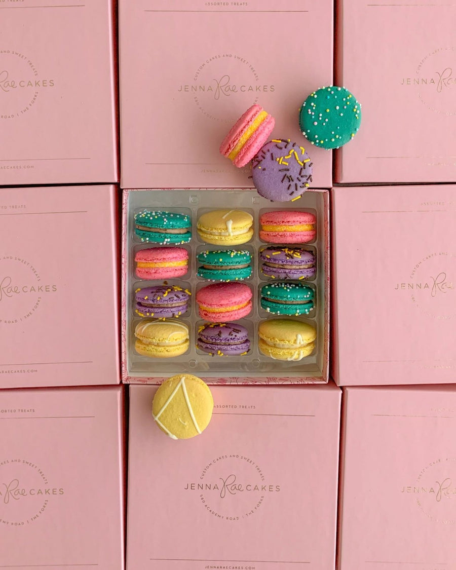A Jenna Rae Cakes Macaron Gift Box featuring yellow, purple, green, and pink macarons sits atop branded pink boxes, with three macarons displayed outside the container.