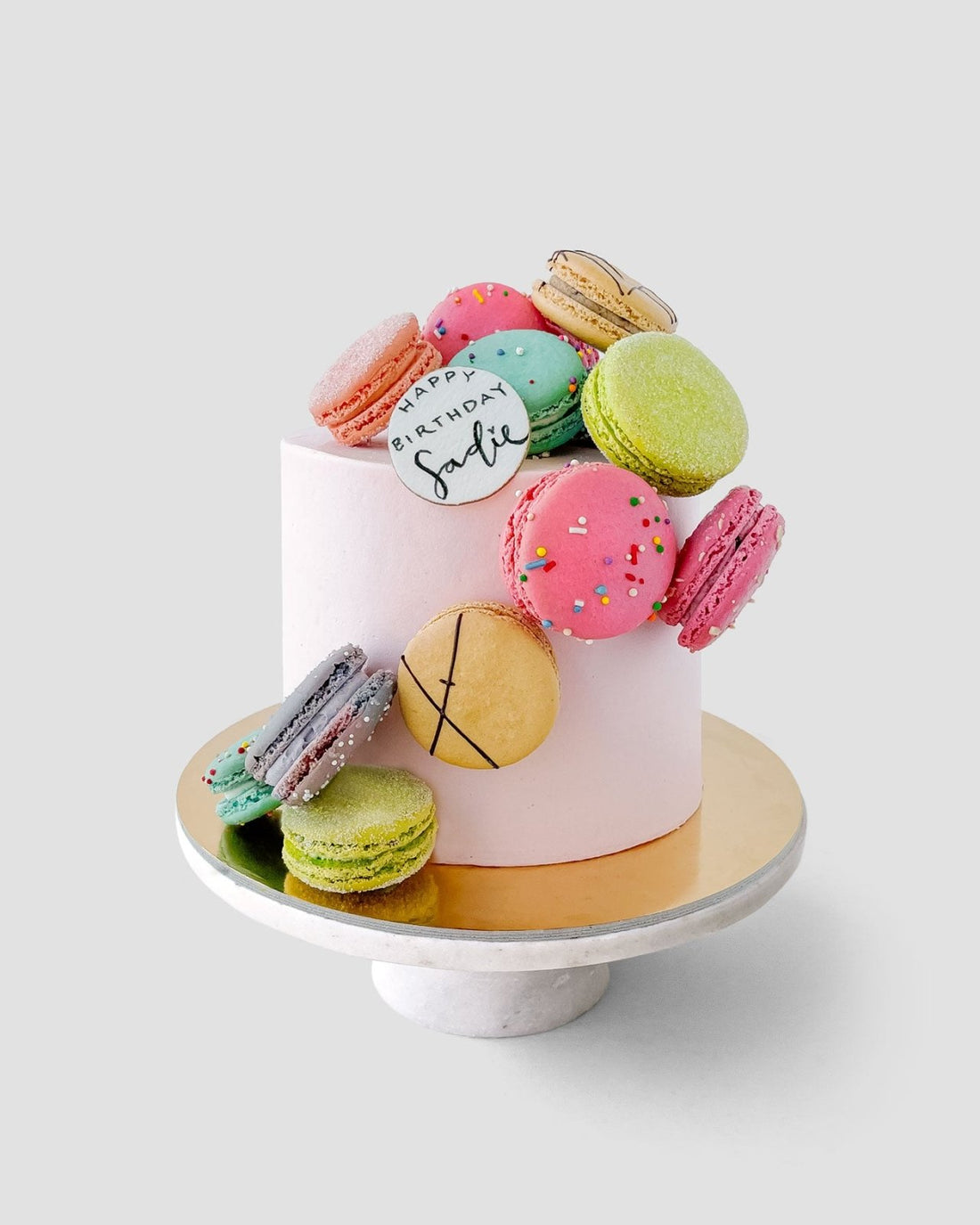 The Macaron Lovers Cake by Jenna Rae Cakes is a white frosted birthday cake adorned with colorful macarons and a white fondant plaque reading Happy Birthday Sadie, presented on a white stand with a gold base.
