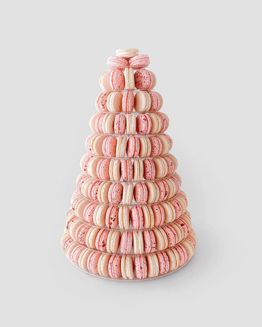 The Macaron Towers by Jenna Rae Cakes feature a tall cone shape with neatly stacked pink and cream macarons arranged in circular tiers, displayed on a light gray background.