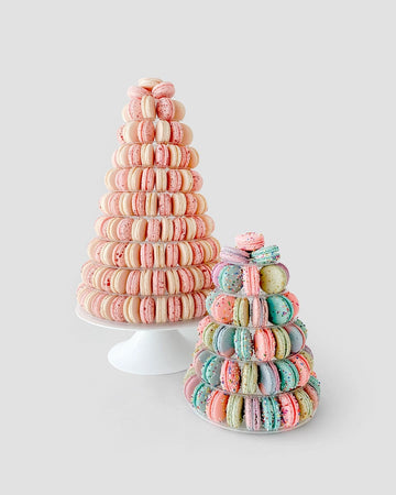 Two Jenna Rae Cakes Macaron Towers are displayed on white stands against a light gray background—one tower features pink macarons, while the other showcases pastel blue, pink, purple, and green macarons.