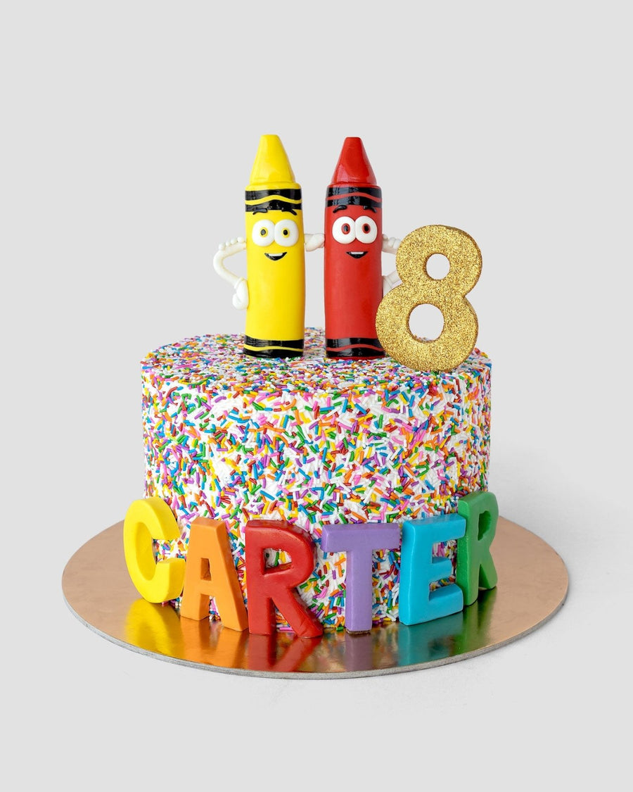 A colorful birthday cake covered in rainbow sprinkles, topped with two crayon figures, a glittery gold number 8, and the name CARTER spelled out in large, colorful letters at the base.