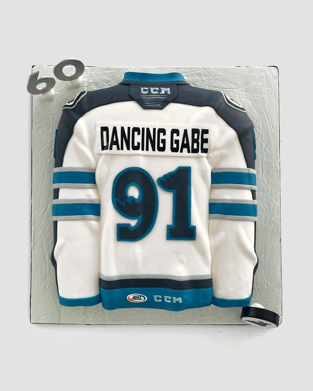 A cake shaped like a white and teal hockey jersey with the name DANCING GABE and the number 91. The cake also features a fondant hockey puck and the number 60 on the corner.