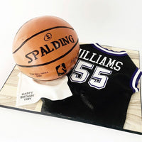A realistic cake shaped like a Spalding basketball and a black basketball jersey with WILLIAMS 55 on it, displayed on a board with a small note that says, Happy Birthday Trev.
