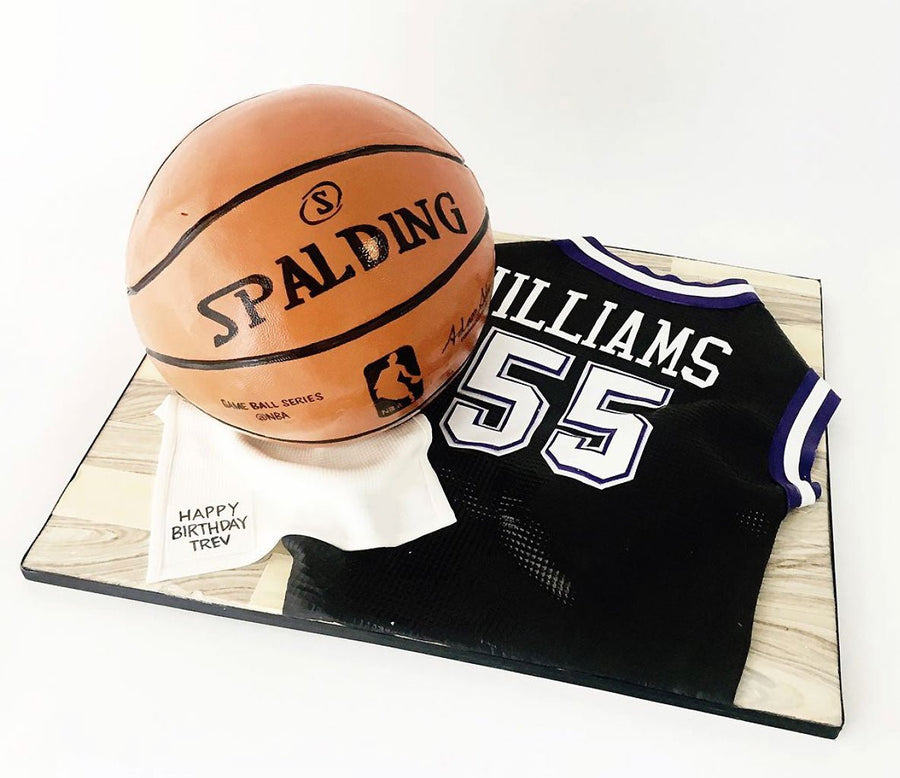 A realistic cake shaped like a Spalding basketball and a black basketball jersey with WILLIAMS 55 on it, displayed on a board with a small note that says, Happy Birthday Trev.