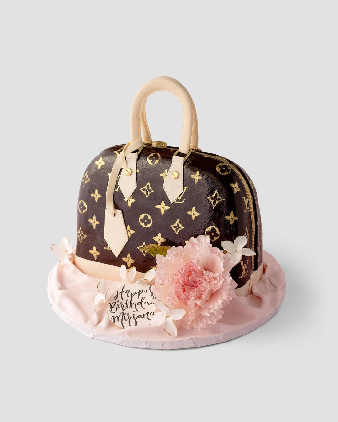 A cake designed to look like a Louis Vuitton handbag, decorated with gold monogram patterns, beige handles, a pink fondant flower, and a “Happy Birthday Mifsara” tag on a pale pink base.