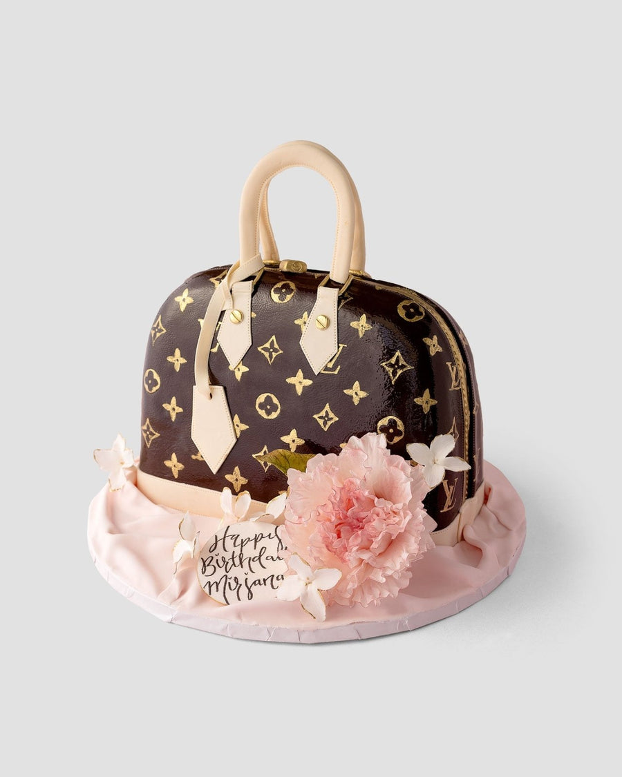 A cake designed to look like a Louis Vuitton handbag, decorated with gold monogram patterns, beige handles, a pink fondant flower, and a “Happy Birthday Mifsara” tag on a pale pink base.