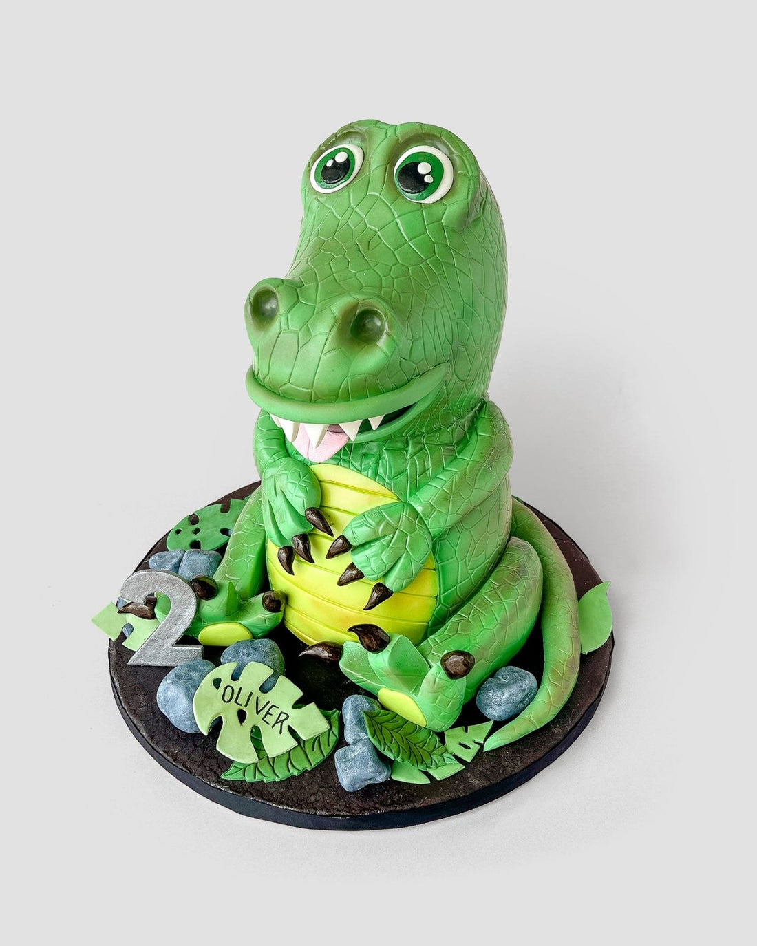 A 3D cake shaped like a smiling green crocodile, sitting upright with big eyes and white teeth, surrounded by edible leaves, rocks, a number 2, and a name plaque reading Oliver.