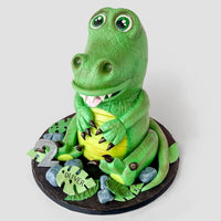 A 3D cake shaped like a smiling green crocodile, sitting upright with big eyes and white teeth, surrounded by edible leaves, rocks, a number 2, and a name plaque reading Oliver.