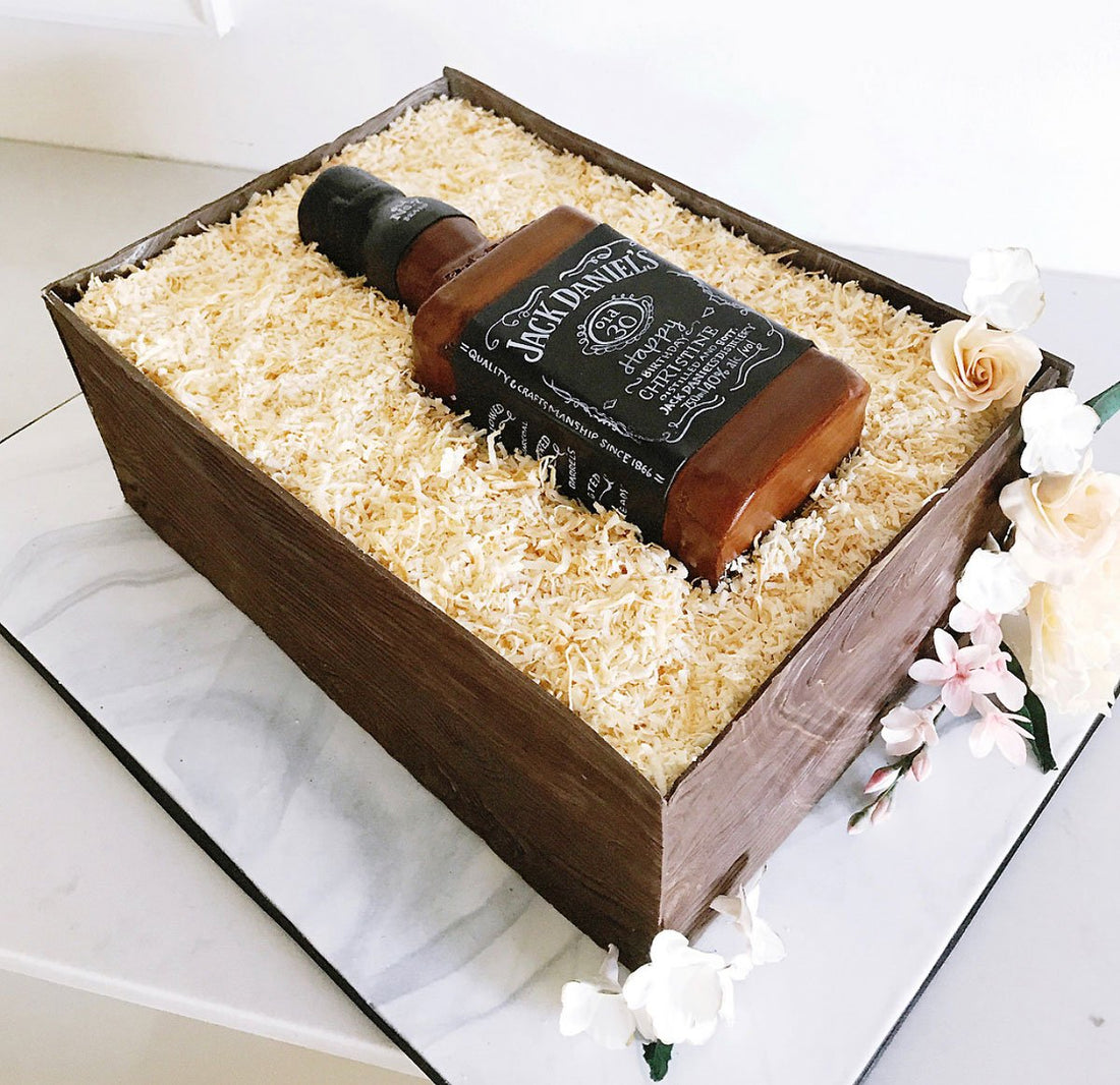 A realistic cake designed to look like a wooden crate filled with shredded paper, containing a bottle resembling Jack Daniel’s whiskey, decorated with white and pale pink flowers.