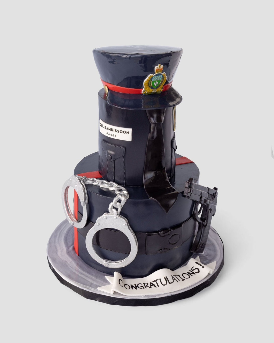 A cake decorated as a police uniform, featuring a hat, badge, handcuffs, walkie-talkie, and a belt. A white banner at the base reads CONGRATULATIONS! The cake is set against a plain background.