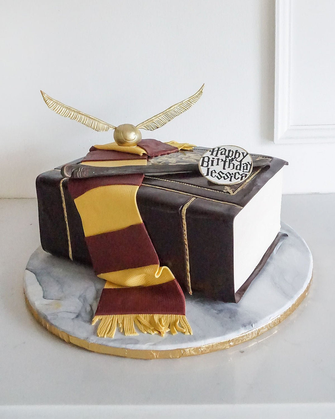 A cake designed to look like a book, decorated with a maroon and gold scarf, a golden snitch, and a plaque that reads Happy Birthday Jessica! on a round marble-patterned board.