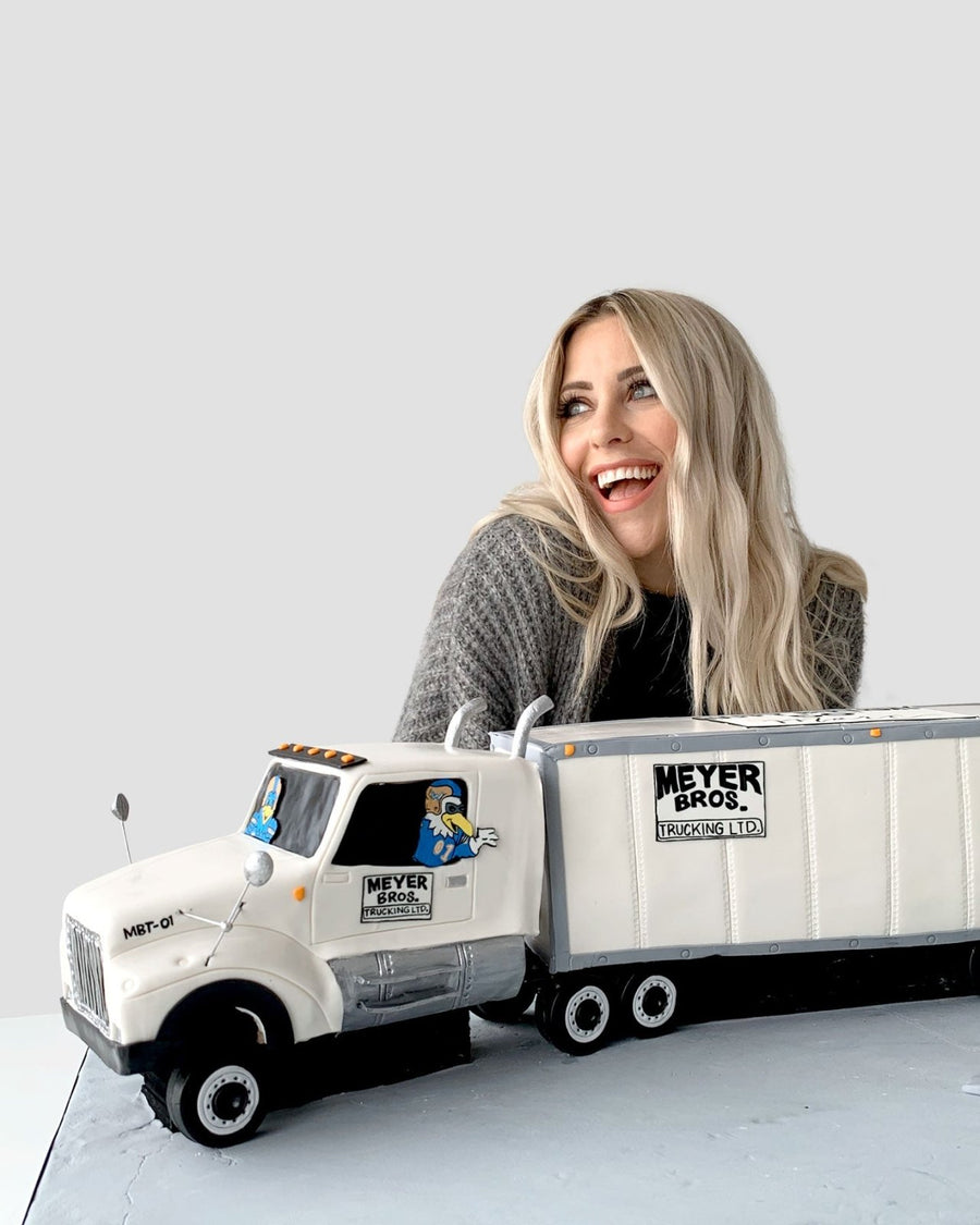 A smiling woman with long blonde hair sits behind a detailed cake shaped like a white Meyer Bros. semi truck and trailer. The background is plain gray.