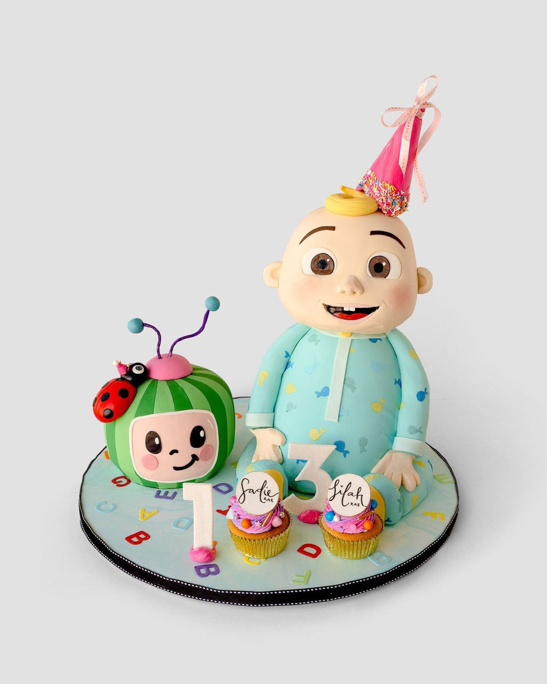 Jenna Rae Cakes’ One-of-a-Kind Cakes: A birthday cake with a baby in blue pajamas and party hat, a green bug with ladybug, number 1 & 2 candles, and two decorated cupcakes on a colorful mat.