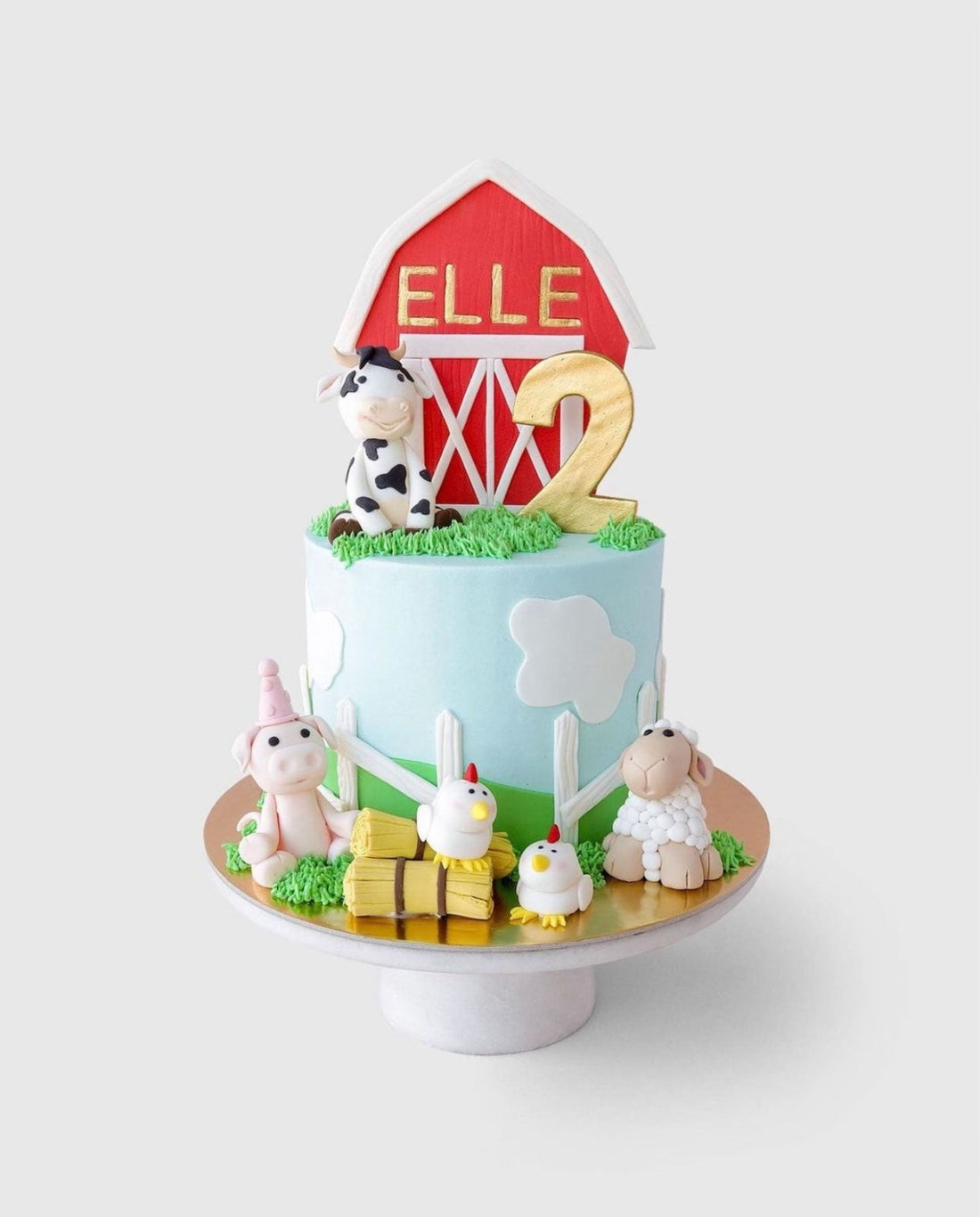 A farm-themed birthday cake with a red barn topper reading ELLE, a gold number 2, and fondant animals including a cow, pig, sheep, and chickens, with clouds and grass decorations.
