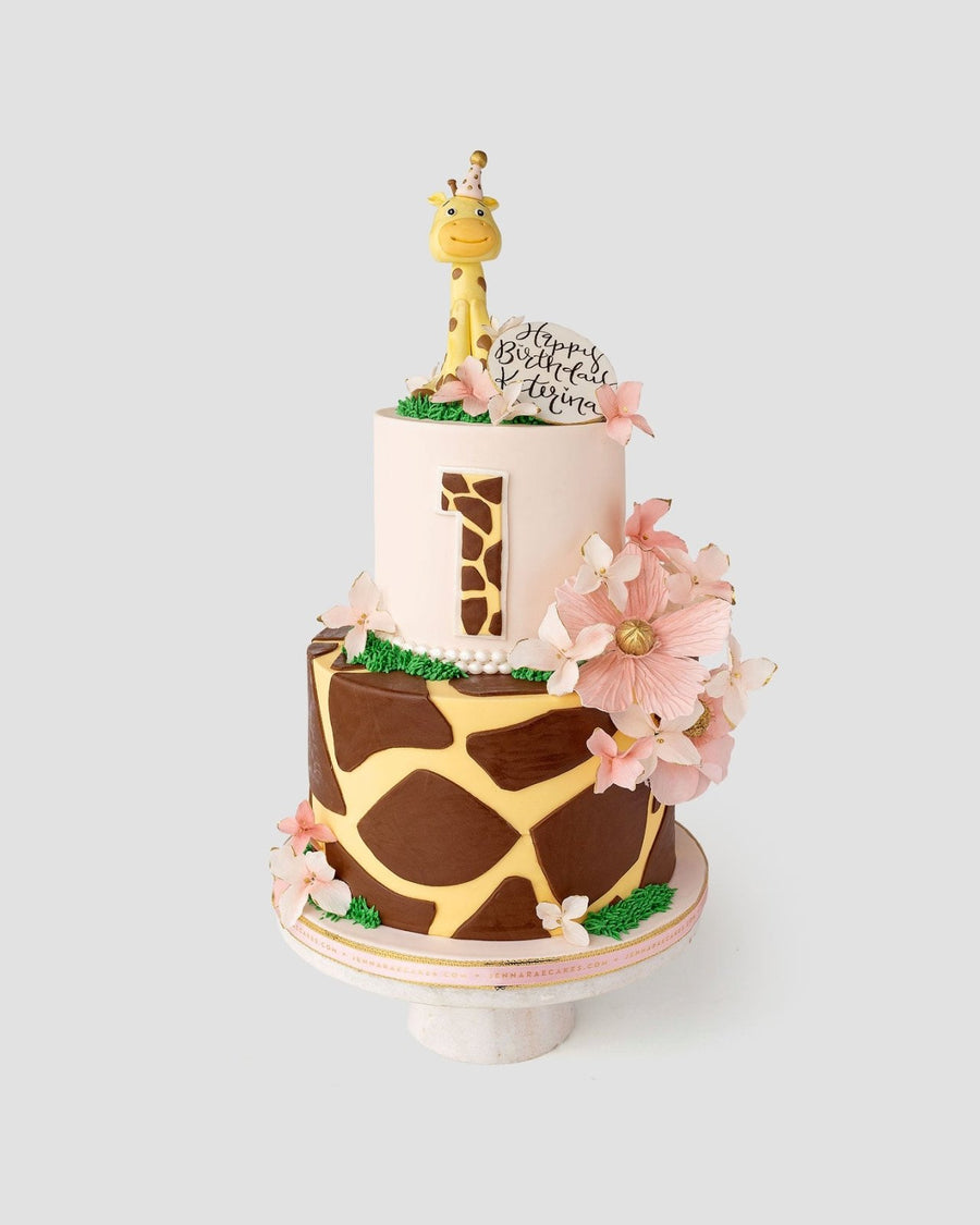 A two-tiered cake with giraffe print patterns, pink flowers, a large number 1, and a giraffe figurine on top, along with a round sign that says Happy Birthday Luna.