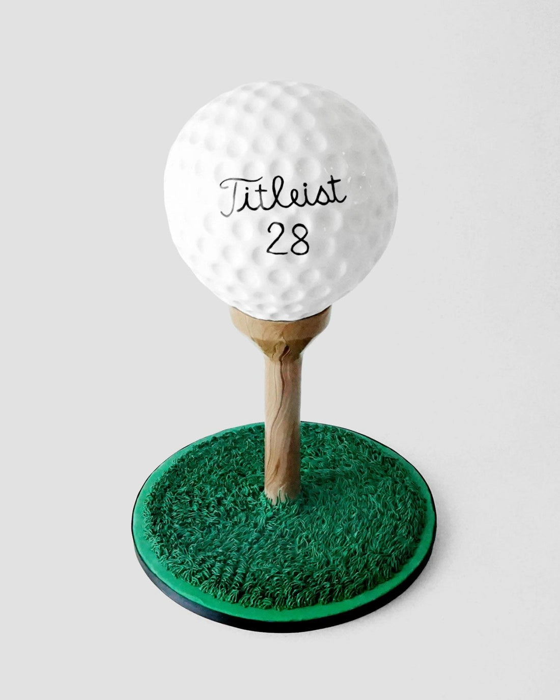 A realistic cake shaped like a golf ball on a wooden tee, set on a circular base decorated to look like green grass, with Titleist 28 written on the golf ball.