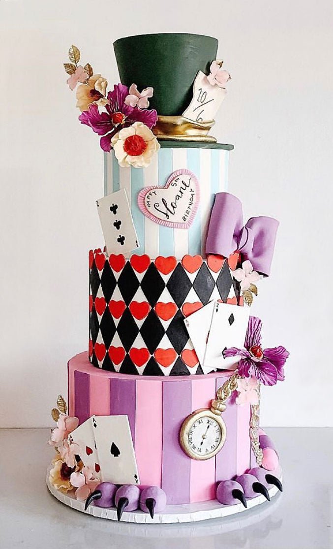 A whimsical, three-tiered cake inspired by Alice in Wonderland, decorated with playing cards, hearts, flowers, a “10/6” hat, a pocket watch, purple bows, stripes, and clawed feet at the base.