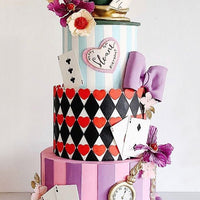 A whimsical, three-tiered cake inspired by Alice in Wonderland, decorated with playing cards, hearts, flowers, a “10/6” hat, a pocket watch, purple bows, stripes, and clawed feet at the base.